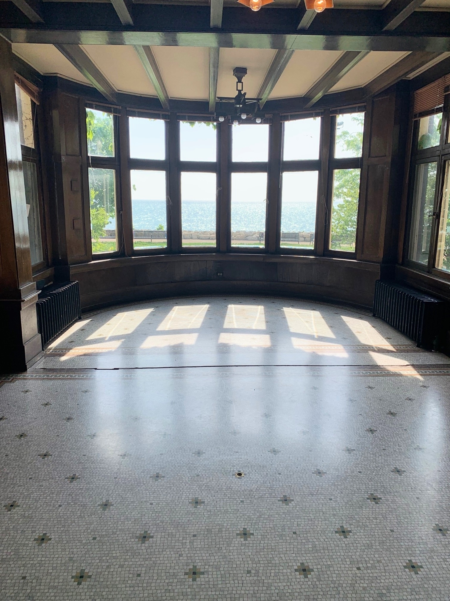 Sunlit room with a curved wall of windows overlooking a lake, mosaic tile floor, and dark wood trim.